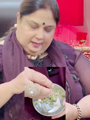 A woman in a purple top is holding a small silver bowl containing green cardamom pods. She is wearing a large, ornate ring and has a black bindi on her forehead. The background includes red and soft warm lighting.