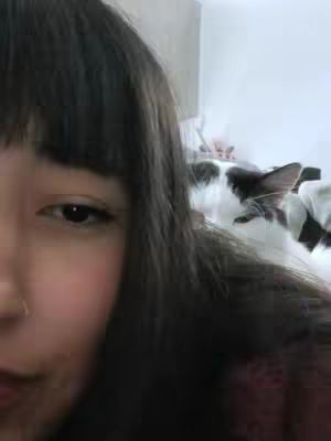 A selfie-style image featuring a woman's face filling a significant portion of the frame on the left. Her dark hair with a fringe partially covers her forehead. The focus is on her eye. To the right and slightly blurred in the background, a black-and-white cat is visible, adding a secondary element to the composition. The overall image is softly lit.