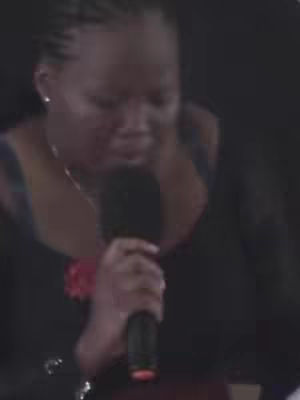 A medium shot shows a woman with dark skin, holding a microphone, looking down with a slight solemn expression. Her hair is styled in braids, and she is wearing a dark outfit and a necklace. The background is dark and indistinct.