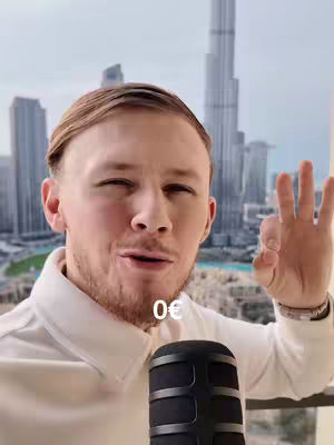 A man in a white shirt is positioned in front of a modern cityscape with tall buildings. He's holding up his hand in an OK sign, and a microphone is in the foreground. The text overlay shows the price "0€".