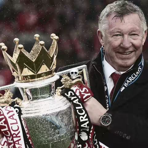 An older man in a black coat and a red tie is holding up a trophy. The trophy has a crown on top and a banner that says "BARCLAYS". The man is smiling. The image has a slightly grainy quality, suggesting it was taken in a slightly blurry environment.
