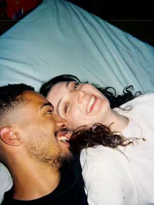 A couple is lying on a bed and looking upwards, faces close together. An adult woman is on the right side and the man on the left. Both are smiling, seemingly mid-laugh. The woman is wearing a white shirt. The bed cover has a slight blue tone. The overall composition is framed from the waist up, capturing the intimacy of the moment.