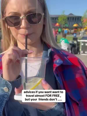 A woman wearing sunglasses is outdoors, holding a clear plastic cup with a straw in her mouth, and there are buildings and greenery in the blurred background. The woman has blonde hair and she is wearing a jean jacket and a red and black plaid overshirt. A text box at the bottom reads, 'advices if you want want to travel almost FOR FREE, but your friends don't.....'