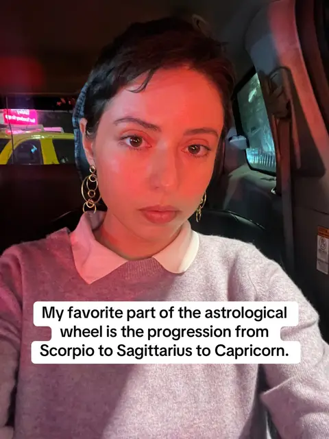A woman with short dark hair and gold hoop earrings sits in the front seat of a car, looking toward the camera. She wears a gray sweater over a white collared shirt. Text overlayed on the image reads: "My favorite part of the astrological wheel is the progression from Scorpio to Sagittarius to Capricorn."
