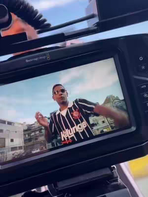 A video is being recorded with a monitor in the foreground, displaying an image of a man wearing sunglasses and a black and white striped jersey with a logo in the top left and the word "kalunga" in red on the front. He is standing on a background of a city scene, with a blue sky.