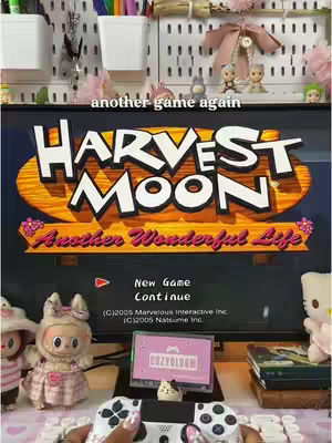 The cover image showcases a gaming setup with a focus on the game "Harvest Moon: Another Wonderful Life" on a monitor. The scene is brightly lit, with a white pegboard background adorned with decorative items. Below the monitor is a desk holding a PS5 controller, with the player's hands visible. The overall impression is of a cozy and inviting gaming environment.