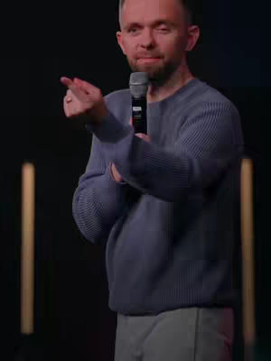 A man wearing a blue sweater and gray pants stands in front of a dark background, holding a microphone and pointing his index finger to the left. The background has vertical yellow lights, The man has short hair, a beard, and a neutral expression.