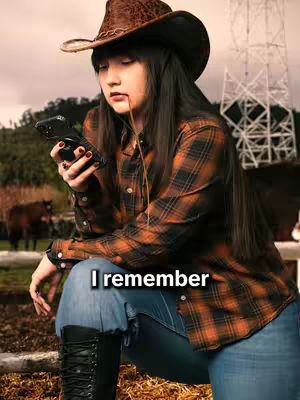 A young woman wearing a cowboy hat and a plaid shirt, holding a phone. She's leaning forward, and a drop of liquid is visible near her mouth. The background shows a blurred outdoor scene with trees and a structure.