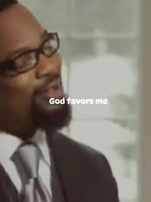 The image shows a man, primarily from the chest up, wearing glasses and a suit with a tie. The text overlay reads 'God favors me'. The background appears to be an indoor setting, likely an office or similar.