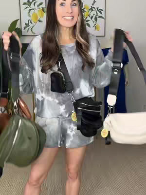 A woman stands smiling, displaying multiple bags of varying styles, with two lemon-themed paintings in the background.