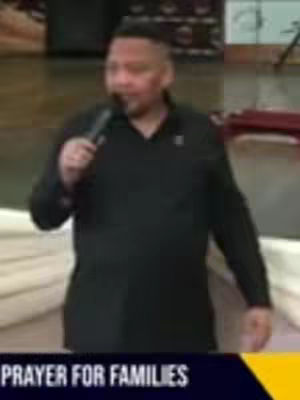 The image shows a man in a black shirt holding a microphone, likely speaking at an event. Below him is a dark banner with the text 'PRAYER FOR FAMILIES' in white.