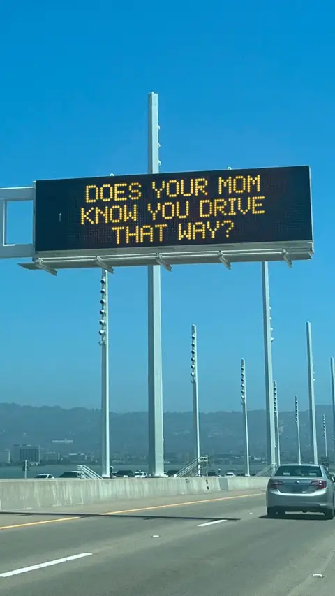 A daytime shot from a car, focusing on a highway with a sign. The sign displays text in yellow, and a grey car is visible in the right lane. Distant mountains and a clear blue sky complete the scene.