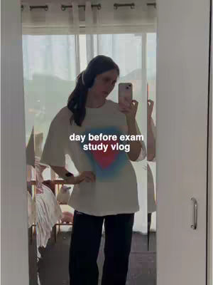 A young woman stands in front of a mirror, taking a selfie. She wears a white t-shirt with a colorful heart design, black pants, and headphones. The background shows a room with a window covered by white curtains.