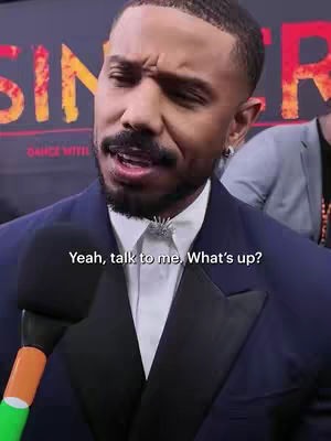 Michael B. Jordan, wearing a dark suit, is speaking into a microphone. The background is blurry but shows the poster of the movie "Sinners". Text overlayed on the image reads "Yeah, talk to me. What's up?"