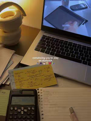 The image shows a desk setup with a laptop, study materials, and a calculator. The laptop screen displays what appears to be a digital drawing or writing. Adjacent to the laptop are sheets of paper, notes, and a calculator. The overall composition conveys a vibe of productivity and cozy study atmosphere.