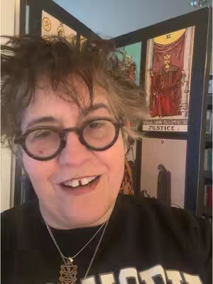 A woman with short, messy, graying hair and round glasses smiles at the camera, revealing a gap in her teeth. She wears a black shirt and a pendant necklace. Behind her is a decorative folding screen with tarot card illustrations and a small bookshelf peeking from the side.
