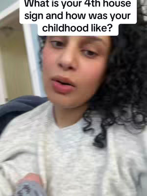A woman with curly black hair and fair skin is the central focus, set against a casual, indoor environment. She wears a light-colored, textured sweater, with a neutral background adding no distinct features or patterns. The composition is dominated by the subject's face, capturing her conversational expression, with the focus on her lips as she speaks. The image has an informal, selfie-like quality, with slightly blurred details and a sense of natural lighting.