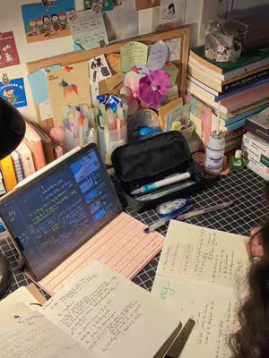 A well-lit, cluttered desk scene. The foreground features open notebooks with handwritten notes, a laptop displaying a blue-themed interface filled with text, and a pink keyboard. A black pencil case, pens, highlighters, and a small stapler are also visible. The background includes a bulletin board with assorted notes and decorations like stickers and photos. The scene conveys an aesthetic study environment.