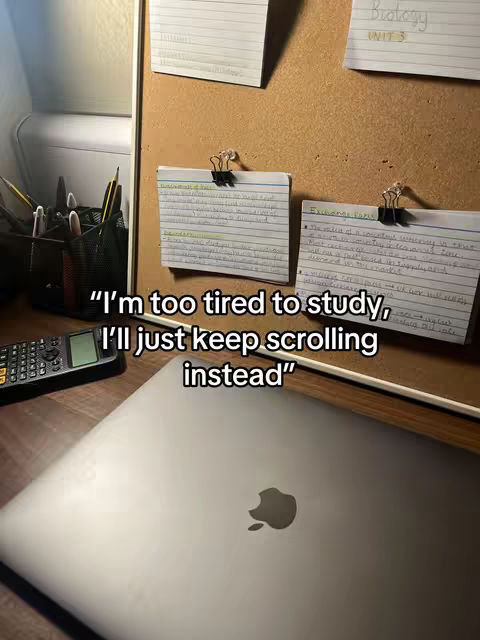 A top-down shot of a desk with study materials, including index cards attached to a corkboard, a calculator, a pen holder, and a laptop. The text overlayed reads, "I'm too tired to study, I'll just keep scrolling instead."