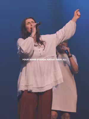 A woman with long dark hair is singing into a microphone on stage with another person in the background. The woman is wearing a white blouse and red pants, and she has her arm raised. The text overlay states, "YOUR NAME STANDS ABOVE THEM ALL."