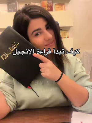 A woman with dark hair, holding a black book with gold lettering, points to it while smiling at the camera. Bookshelves are visible in the background.
