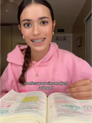 A young woman with a pink hoodie and braces is looking directly at the camera while sitting in front of an open Bible. Text overlayed at the bottom reads, 'confess your sins and just ask god'.