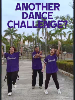 The image features three individuals in an outdoor setting, posing dynamically. They are all wearing purple t-shirts with the word "Revival" in white script across the chest. The background includes lush green palm trees and a concrete walkway. The text overlay reads "ANOTHER DANCE CHALLENGE?" in a bold, dark purple font.