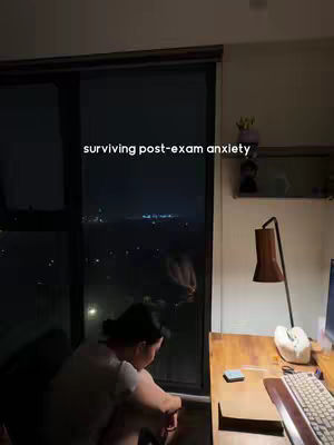 A dimly lit interior shot shows a person looking out a large window at a cityscape at night. Text overlayed on the image reads "surviving post-exam anxiety". A desk with a computer is visible nearby.