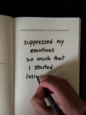 An open notebook page with handwritten text, a hand holding a black pen is writing on the page. The text reads: "what's one thing that drains you that you keep doing? suppressed my emotions so much that i started losing". The background is dark.