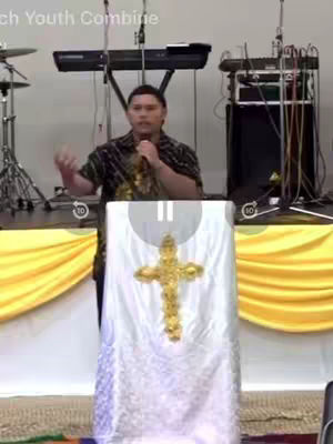 A man, likely a preacher, stands behind a podium with a gold cross. He is gesturing with his right hand while holding a microphone. The setting appears to be a church, with sound equipment and instruments visible in the background and curtains.