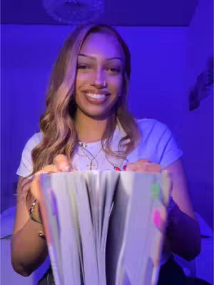 A woman with long blonde hair is holding open a book, looking at the camera and smiling. The background is lit with purple light, and she is wearing a white t-shirt and a necklace.