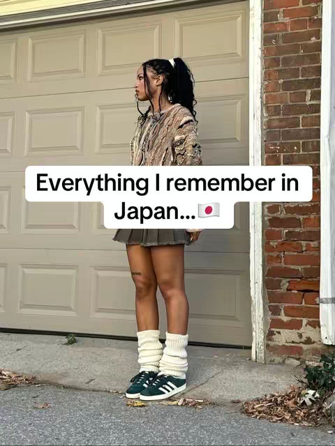 A woman stands outside, facing left, with a garage door and a brick wall in the background. She wears a patterned long-sleeved shirt, a pleated skirt, leg warmers, and sneakers. The image has a neutral color palette with the text overlayed.