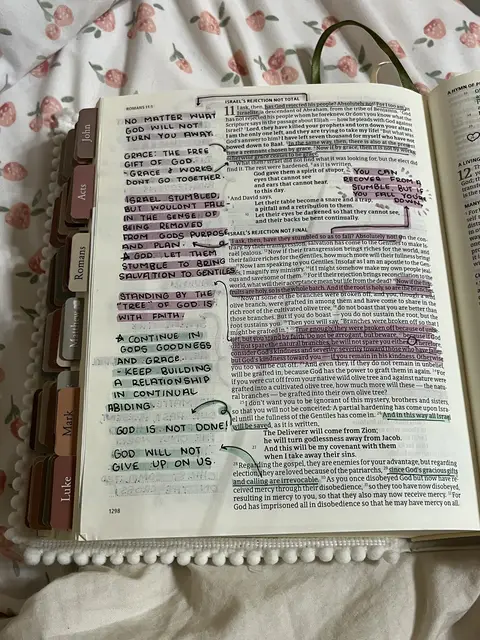 An open Bible with highlighted and annotated text. The pages are open to Romans 11, with multiple areas highlighted in pink and yellow, and handwritten notes in black. The backdrop is a floral patterned fabric in pinks and whites. Tabs for books in the Bible are visible along the side.