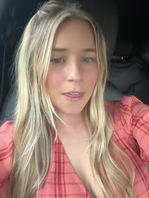 A selfie of a woman with blonde hair and a pink plaid shirt, her tongue slightly out, with a focus on her face. The lighting is soft, and the background suggests a car interior.