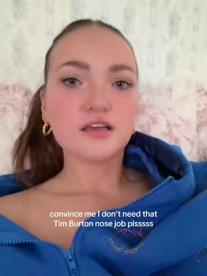 A woman with light skin and light brown hair is centered in the image, looking at the camera. She has a slight smile. Her hair is styled in a low ponytail, and she wears small gold hoop earrings. She is wearing a blue zip-up jacket. The background is a soft pattern, likely a floral design.