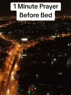 An aerial shot at night showcasing a cityscape illuminated by warm-toned lights. A major road with traffic extends towards the bottom, while buildings and streets form the background. The sky is dark, with the glow of city lights creating a vibrant night scene. The text overlay reads: "1 Minute Prayer Before Bed".