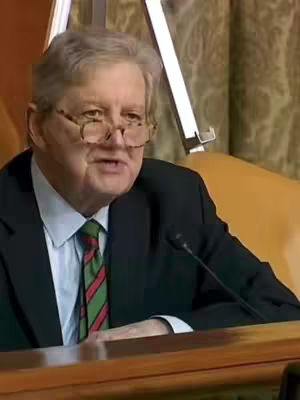 The cover image shows a middle-aged man with gray hair and glasses, seated at a table. He is wearing a dark suit, a light blue shirt, and a tie with green and red stripes. A microphone is positioned in front of him, and an easel-like structure is visible in the background. The lighting appears to be from overhead, illuminating the man's face and upper body. The composition is framed from the chest up, with the man speaking and looking off to the side. The color palette is composed of muted tones typical of a legislative setting.
