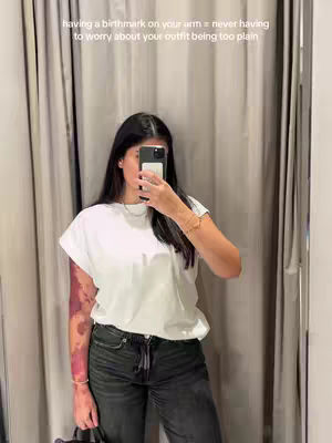 A person stands in front of a curtain, partially obscuring their face with a phone. They wear a white t-shirt and dark jeans, revealing a birthmark on their arm. The background features neutral-toned curtains.