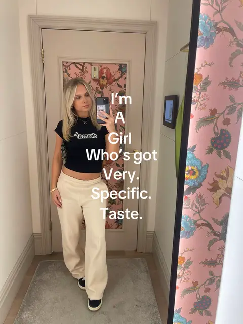 A woman is in a dressing room, taking a selfie in front of a mirror. She's wearing a black top and off-white pants. Text overlays are on the image, and the background features decorative wallpaper.