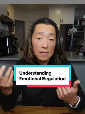 A woman with long brown hair is centered in the frame, set in a well-lit kitchen. She is wearing a black top and her hands are up, framing a text overlay reading 'Understanding Emotional Regulation.' Behind her are white cabinets and stainless steel appliances. The background is slightly blurred, focusing attention on her face and the overlaid text.