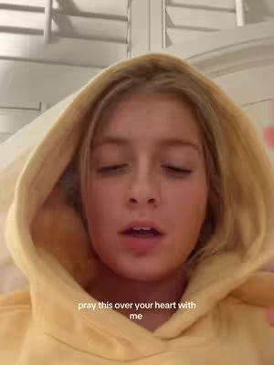 A close-up shot features a well-lit young woman with blonde hair gently framing her face. She's wearing a light yellow hoodie, and the background has white blinds. The image is focused on her face, highlighting her facial features and expression.