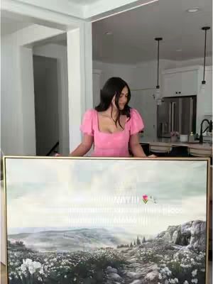 A woman in a pink dress poses behind a framed canvas. The canvas displays a landscape painting with a prominent path and white flowers, set against a light, atmospheric background. The background includes a kitchen setting with white cabinets and pendant lighting.