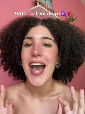 A selfie-style image featuring a young woman with curly dark hair and rosy cheeks positioned in the center of the frame. She has an open mouth expression and her eyes are looking at the camera. Text is overlaid at the top, with a pink wall background
