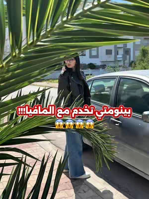 A woman stands near a car, partially obscured by large green leaves in the foreground. The background shows a street and buildings. The woman is wearing a jacket and jeans, and text in Arabic is overlaid on the image.