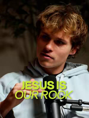 A young man with light brown hair is centered in the frame, wearing a light blue hoodie. He is posed towards the right and pointing his right hand up, a microphone in front of him. The background is softly blurred with neutral tones. Overlayed, in bright yellow-green, is the text 'JESUS IS OUR ROCK'.