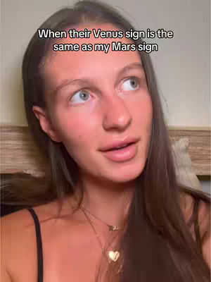 A close-up shot of a woman with long brown hair and fair skin, looking slightly upwards with a contemplative expression. She wears a black top, and a gold necklace with a heart pendant. The background is a blurred, neutral tone, likely of a wall or headboard. Text overlayed on the image reads, "When their Venus sign is the same as my Mars sign."