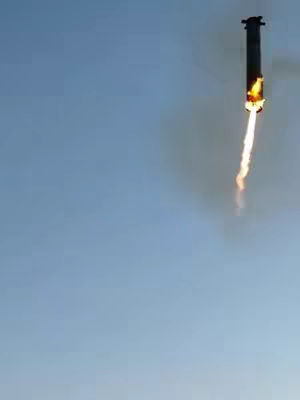 A rocket booster in mid-air, with a trail of fire and smoke extending downward. The background is a clear blue sky.