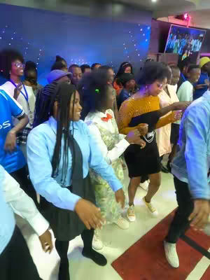 A group of people are dancing in a brightly lit hall, some are holding drinks.