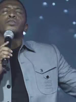 The cover image shows a medium shot of a Black man singing into a microphone. He is wearing a light gray jacket over a black shirt. His eyes are closed, and he appears to be in a performance setting, with a dark background and some blurry lights.