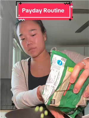 A woman is shown in a kitchen setting, holding a green bag filled with what appears to be green vegetables. She is pouring the contents into a dark-colored pan. Above her, a red bar with white text reads
"Payday Routine."